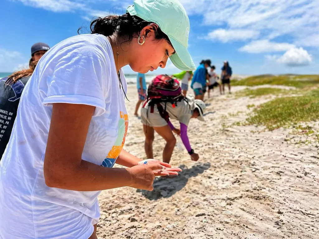 A volunteer collects microplastics by hand on a beach in Galápagos while others work in the background.

Title